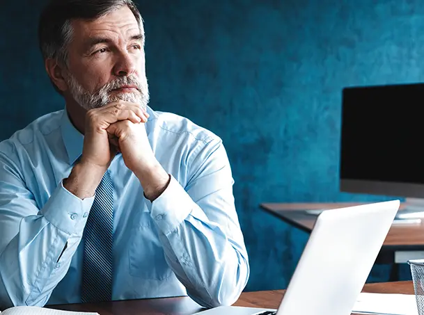 Senior executive in a blue shirt sitting at a desk with a laptop, reflecting on business strategy.