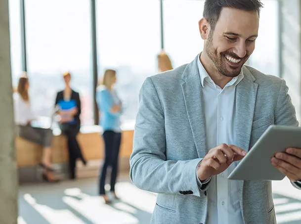 Business professional smiling while using a tablet, with colleagues collaborating in the background.