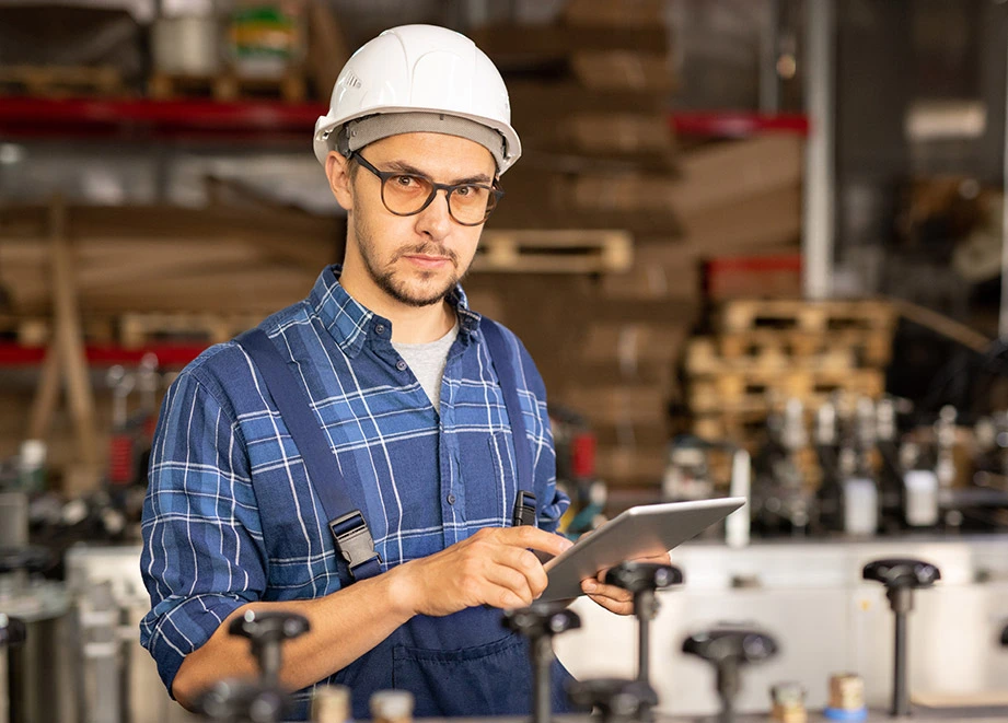 Worker in safety helmet and glasses using a tablet in a factory setting.